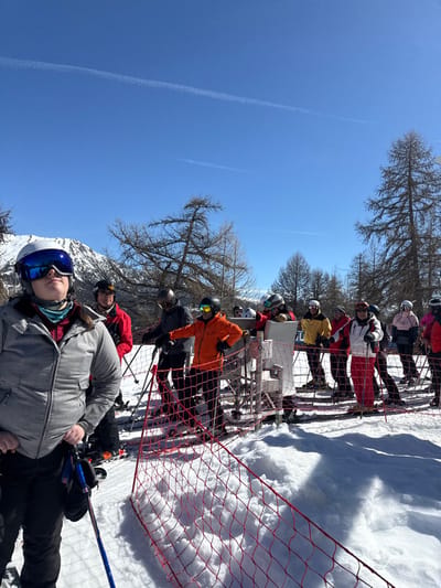 The image captures a group of approximately ten to twelve skiers queuing on a snow-covered mountain slope at a ski resort in Saint-Chaffrey, France. The scene is set under a clear, bright blue sky with faint white contrails. In the foreground, a red safety net with square mesh acts as a barrier. Behind it, skiers, dressed in diverse winter attire including helmets, goggles, and brightly colored ski jackets (grey, red, orange, yellow, white, pink), stand with their skis on the snow and ski poles in hand. A white ski lift gate or turnstile structure is visible mid-ground on the right, towards which the skiers are oriented. Bare, deciduous trees, characteristic of alpine environments, line the mid-ground and background, with snow-capped mountains visible on the left horizon. One individual in the immediate foreground on the left wears a grey jacket, blue mirrored goggles, and a white helmet, looking upwards.