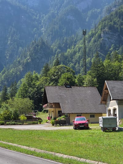 The image depicts a rural scene in Landl, Austria, featuring a cluster of houses nestled at the base of a steep, forested mountain. In the foreground, a paved road runs along the bottom left of the frame, bordered by a grassy verge and a stone curb. A well-maintained lawn stretches across the middle ground, dotted with small yellow wildflowers.
A bright red car, identified by a license plate that appears to read "SB 822 AE," is parked on a gravel area. Beside the car, a small, light-colored utility trailer with the license plate "LI 119 DZ" is hitched to an unseen vehicle or parked on its own.
Dominating the middle ground are two houses. The larger house on the left has a dark, pitched roof with diamond-shaped tiles, a yellow facade, and a wooden balcony adorned with pink flowers. The smaller house on the right is partially visible, with a similar pitched roof and a concrete block facade.
Behind the houses, a wooden fence runs horizontally, separating the lawn from a sloped area with more vegetation and a stack of firewood. Beyond this, the landscape rises sharply, covered in a dense forest of coniferous and deciduous trees. The mountain slopes are steep and rugged, with rocky outcrops visible in places. In the background, a tall, dark metal telecommunications tower stands prominently against the green foliage. The sky above is clear and bright, suggesting a sunny day. The overall impression is one of a serene, mountainous village setting.