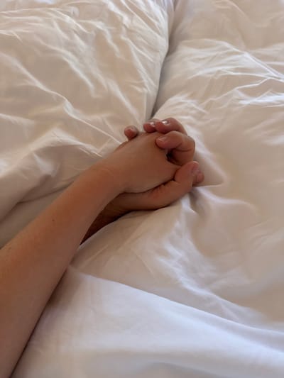 The image shows two hands intertwined on a white, crumpled bedsheet. The hands appear to be of different skin tones, suggesting they belong to two different individuals. The lighting is soft, casting gentle shadows on the fabric, highlighting the textures of the sheets. The overall impression is one of intimacy and connection. The location is described as Kovačka Dolina, Montenegro.