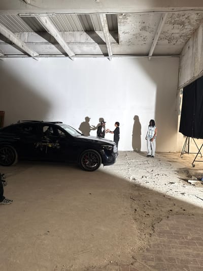 The image shows a film set or photoshoot in an interior location. The location features a white wall on one side and a wall with peeling paint and structural elements on the other. Two people stand near a black car, one leaning over the hood, and another stands further away. A black backdrop hangs from lighting stands in the background. The floor is concrete with debris and cracks. Based on the architectural style and apparent disrepair, the location may be a former industrial space or warehouse. The location is Ivry-sur-Seine, France.