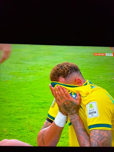 The image captures a close-up, medium-angle view of a male soccer player, likely Neymar Jr., in a yellow jersey with blue and green accents, on a vibrant green soccer field. The player is covering his face with both hands, and a yellow shirt is draped over his head, obscuring his features. He is wearing a white wristband on his left arm, and his right arm displays intricate tattoos. A patch on his jersey reads "Unite For Education FIFA." In the background, a small red and white graphic displays "НА ЖИВО Б-Н-Т," indicating a live broadcast by BNT, a Bulgarian National Television channel. The scene appears to be from a professional soccer match. There is no information to determine the location within the city of Ognianovo, Bulgaria.