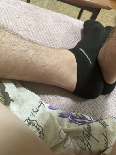 The image shows a close-up view of a person's legs and feet, wearing black socks. The person's legs are hairy. The background consists of a textured pink surface, possibly a blanket or rug. A patterned fabric with what appears to be writing and illustrations is draped in the foreground. The scene is indoors, and there is no visible indication of the city of Corlăteni, Moldova.