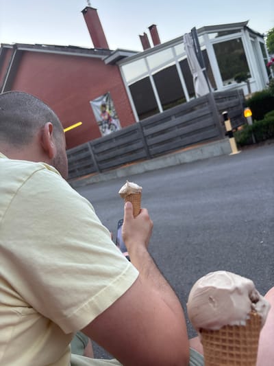 The image depicts a close-up, over-the-shoulder view of a person's head and arm, holding an ice cream cone. The person is wearing a pale yellow t-shirt. Another ice cream cone is visible in the foreground, held by a person's hand. In the background, a red brick building with a distinctive chimney and a sunroom is present. A wooden fence runs along the property line, and beyond that, a road or paved area is visible. The overall setting appears to be an outdoor area, possibly a restaurant or cafe, in Zutendaal, Belgium.