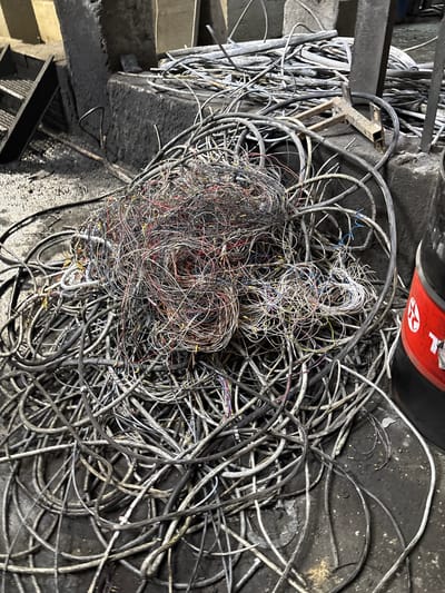 This overhead shot depicts a substantial, chaotic pile of electrical wires and cables, discarded or stored on a dark, dirty ground. The scene appears to be an industrial setting, likely a scrap yard, recycling facility, or workshop, located in Mirkovo, Bulgaria.
The primary subject is a dense entanglement of wires. In the center, a large mass of thin, multi-colored wires (featuring prominent reds, blues, yellows, greens, and whites) forms a tight, disorganized knot. Surrounding this, and extending across the frame, are numerous thicker cables and conduits, predominantly grey, black, and off-white, lying crisscrossed and coiled on the ground. The ground itself is rough, dark, and appears to be concrete or asphalt, stained with grime and debris.
To the upper left, some metal structural elements, possibly beams or racks, are visible, partially obscured by more bundles of cables. A segment of a red cylindrical object, likely a barrel or container, with a white star-like emblem, is present in the upper-middle section of the image. No people or activities are visible, suggesting a static collection of materials rather than an ongoing process. The lighting is uniform, hinting at an indoor or covered environment, and no specific time of day or weather conditions can be discerned. There is no legible text on any of the visible objects beyond the indistinct white symbol on the red container.