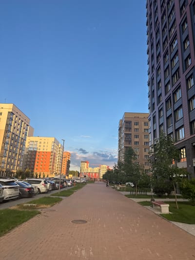 This is an outdoor, eye-level shot of a modern residential area in Koltsovo, Russia. The scene is dominated by a wide, reddish-brown brick paved walkway that extends into the distance, lined with green grass. To the left of the walkway is a row of parked cars, including white SUVs and sedans. Beyond the cars, several multi-story apartment buildings are visible, varying in color and architectural style, with a clear blue sky overhead. To the right of the walkway, a dark, modern apartment building stands tall, featuring rows of windows. Several trees with green foliage are planted between the walkway and the buildings, and a park bench is visible on the right side. In the distance, more apartment buildings and some construction cranes are present. A few indistinct figures are walking on the walkway. The lighting suggests it is late afternoon or early evening, with long shadows cast by the buildings.