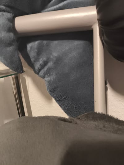 This media file shows a close-up view of a white, tubular metal frame, possibly part of a piece of furniture or a structure. The frame is partially covered by soft, fuzzy fabric, predominantly in shades of grey and dark blue. The fabric has a plush texture, suggesting it might be part of a blanket, sofa, or pet accessory. In the background, a white, textured wall is visible, illuminated by what appears to be artificial light, indicating an indoor setting. On the left side, a metallic rod or leg of another object is partially visible, along with a hint of a glass surface. The composition is abstract, focusing on textures and forms rather than a clear depiction of a specific object or scene. There is no visible text. The lighting suggests it is either daytime with artificial lighting or nighttime. The context of Karlsruhe, Germany, does not provide any direct visual cues within the image itself.