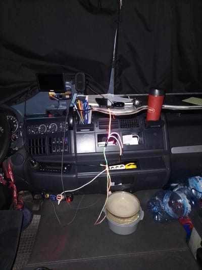The image depicts the interior of a truck cab, likely in Bad Honnef, Germany, given the context. The scene is dimly lit, with black curtains drawn across the upper part of the frame, obscuring the view outside. The dashboard is cluttered with various items.

Prominently displayed on the dashboard are a small screen, possibly a GPS or camera monitor, mounted above a car charger with a digital display showing "25.3". There are also several electrical outlets and a red USB adapter. A black mesh cup holder is filled with pens and markers. To the right, a red insulated travel mug is positioned in a holder.

In the lower section of the cab, on the floor, a grey plastic bucket contains a lighter-colored bowl, with what appears to be a yellow object and some cords draped over it. Several large plastic bottles of water, labeled "Polaris 5L", are also on the floor. A dark-colored car floor mat is visible, along with a blue sandal. The overall impression is one of a lived-in space, possibly a driver's resting area.