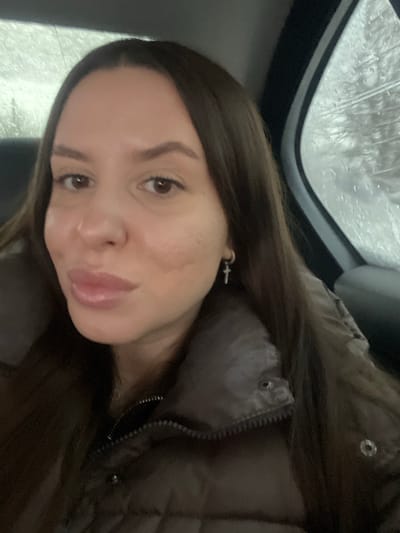 The image displays a close-up, frontal view of a female subject, appearing to be a young adult, situated within the interior of a vehicle. Her long, straight, dark brown hair is parted centrally. She has brown eyes, a fair complexion with some textural variations on her cheeks, and full, glossed lips. She is wearing a dark brown puffer jacket with a visible zipper and snap fasteners. A silver-toned hoop earring with a small cross pendant is present on her right earlobe. The subject is looking directly at the camera with a slight, closed-mouth smile. Through the vehicle's right-side window, an out-of-focus exterior environment is visible, characterized by water droplets on the glass, indicating precipitation. The background outside the vehicle appears uniformly light gray/white, suggestive of an overcast sky or snowy conditions. No specific geographical markers or landmarks are present to confirm a location in Vysokovo, Russia.