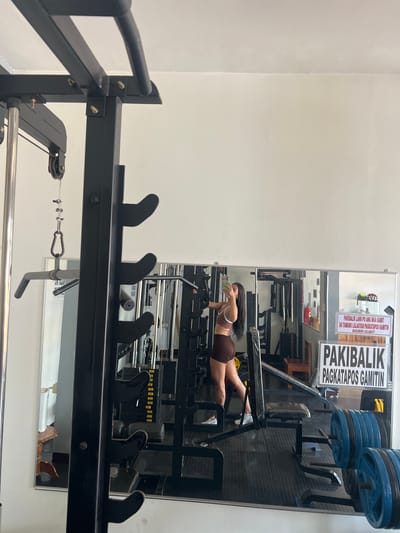 A woman is taking a selfie in a gym. She is wearing a brown sports bra and shorts. The gym is equipped with various exercise machines and weights. There are signs on the wall in the background with text that appears to be in Filipino. The location is specified as Cuyo, Philippines.