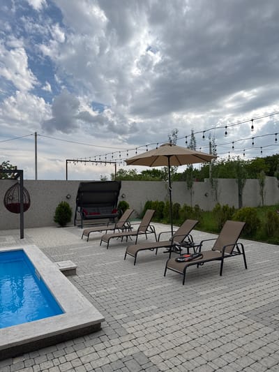 The image depicts an outdoor patio area adjacent to a swimming pool, likely located in Mughni, Armenia. The patio is paved with light gray interlocking stones. Several lounge chairs made of dark brown fabric and metal frames are arranged on the patio, with one chair in the foreground displaying a pair of rackets. A large, light brown patio umbrella stands open near the lounge chairs. In the background, a dark gray swing set with red cushions is visible against a textured gray wall. To the left, a dark circular hanging chair with a red cushion is suspended from a metal frame. A portion of a bright blue swimming pool with a light gray stone border is visible in the lower left corner of the frame. String lights are suspended across the upper portion of the image, and a dense green hedge borders the far side of the patio. The sky is overcast with dramatic, gray clouds.
