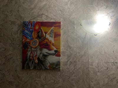 A canvas painting of a fox wearing a headdress is displayed on a textured wall. The fox is depicted in profile, facing to the right, with a colorful headdress adorned with feathers and beads. The background of the painting is a sunset or sunrise with warm colors. To the right of the painting, a wall-mounted light fixture with a white, translucent shade is illuminated, casting a bright light on the wall. The wall itself has a textured pattern resembling abstract swirls. The scene appears to be an interior setting. The location is identified as Sukhoy Ruchey, Russia.
