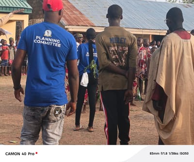 Here is a detailed description of the image:
The image shows a group of people standing in an outdoor area, likely in Langtang, Nigeria. In the foreground, four people are facing away from the camera. The first person on the left is wearing a blue t-shirt with "PLANNING COMMITTEE" printed on the back, along with light-colored jeans that have a logo on the back pocket. The second person from the left is wearing a blue shirt with what appears to be "PLANNING COMMITTEE" partially visible, paired with black leggings and sandals. They have long hair in a ponytail and are holding green foliage. The third individual is wearing an olive-green shirt with "MOSLEY" and the number "57" printed on the back, along with black pants that have a red and white stripe. On the rightmost side, a person is draped in a light beige garment and is wearing glasses.
Behind these four individuals, a crowd of people, including children and adults, is gathered, and there is a low building with a corrugated metal roof in the background. The ground is covered in dirt. The image also features camera information at the bottom, indicating the camera model is a "CAMON 40 Pro" and providing the technical details of the shot (83mm f/1.8 1/386s ISO50).
