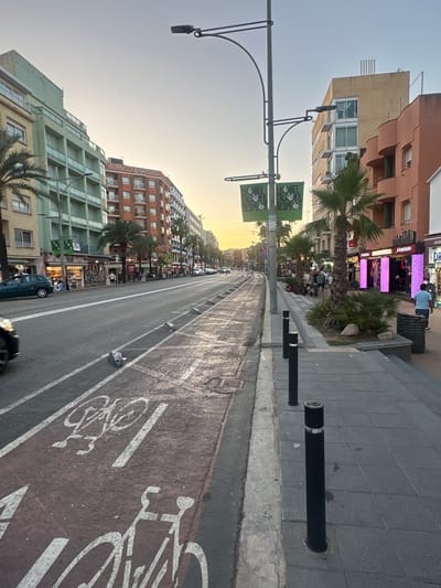 The image shows a street scene in Lloret de Mar, Spain. A street with a bike lane painted in red and white, marked with bicycle symbols, is visible in the foreground. Several parked cars and a moving car are on the road. On the right side of the street, there is a sidewalk with black bollards and a few palm trees. Shops with illuminated displays line the sidewalk. In the background, multi-story buildings with balconies and windows are visible. The sky is a pale orange, suggesting either sunrise or sunset. Several people are walking on the sidewalk.