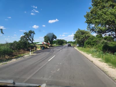 The image depicts a road scene in Krushovitsa, Bulgaria, on a clear, sunny day with scattered white clouds. The perspective is from within a vehicle, looking forward along a two-lane asphalt road that cuts through a rural landscape. Lush green trees and bushes line both sides of the road, with some drier, brush-like vegetation on the left embankment.

On the left side of the road, near a bend, there is a small roadside stall or market setup. A green and white striped canopy covers a display area with what appear to be goods for sale, possibly local produce or crafts. Two Bulgarian flags are prominently displayed on poles near the stall, fluttering in the breeze. A person in dark clothing is standing near the stall, seemingly attending to customers or the setup. A small table or counter is visible under the canopy, and some teal-colored crates or containers are stacked nearby. The road surface in front of the stall shows signs of recent work, with patches of darker, possibly freshly laid asphalt.

Further down the road, a dark-colored car is visible, moving in the same direction as the viewer. To the right of the road, the vegetation is dense and green, with tall grass growing along the shoulder. White and red striped road markers are visible at intervals along the roadside. The overall impression is a snapshot of a typical day in a Bulgarian rural area, possibly capturing a moment of local commerce and daily travel. The time of day appears to be midday, given the bright sunlight and clear sky. There is no visible text in the image.
