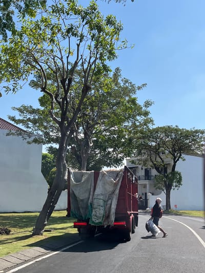 In Entalsewu, Indonesia, a person is crossing a paved road, carrying two large white plastic bags. The person is wearing a dark jacket, shorts, and a light-colored head covering, and appears to be wearing sandals. They are walking in front of a large, dark red truck that is parked or moving slowly on the road. The truck has its rear brake lights illuminated and is covered with large, light-colored tarps. In the background, there are white buildings with balconies and tiled roofs, and several large trees with green foliage. The ground is covered with a neatly manicured lawn. The sky is clear and blue.