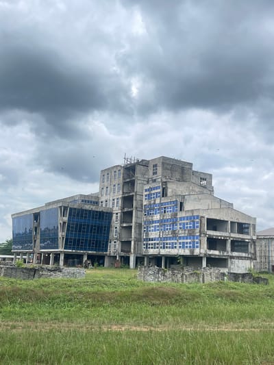 The image shows an unfinished concrete building in Abraka, Nigeria. The building has several stories with large window openings. Some windows are filled with reflective blue glass panes. Overgrown green vegetation surrounds the base of the building, and a cloudy, overcast sky fills the background. There are no people visible in the image.
