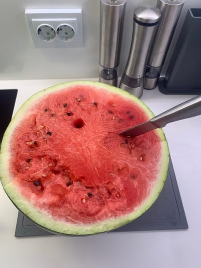 A close-up, eye-level shot shows the interior of a halved watermelon on a gray cutting board. The watermelon flesh is a vibrant red, speckled with numerous black seeds. A silver knife is inserted into the watermelon, angled downwards. In the background, out of focus, are two stainless steel salt and pepper grinders and a white electrical outlet with two circular sockets. The overall setting appears to be a kitchen. The image was captured in Bugry, Russia.