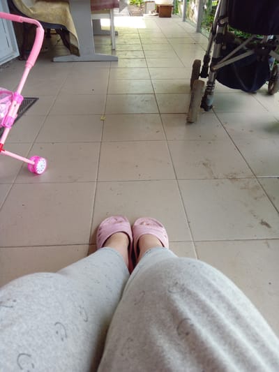 The image depicts a tiled porch with two pink rubber flip-flops on the feet of an individual wearing gray sweatpants with a subtle smiley face pattern. To the left, a pink toy stroller with white wheels is visible, positioned on a dark doormat. In the background, a portion of a wheelchair with a black seat is visible. Further back, a table covered with a patterned cloth is partially seen, with the legs of chairs underneath. The porch appears to be outdoors, with sunlight casting shadows on the tiled floor. The scene is set on a porch in Kitskany, Moldova.