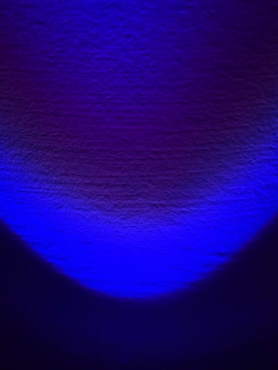 The image depicts a heavily textured wall surface, illuminated by a distinct light source, located in the City of Derby, United Kingdom. A prominent, arc-shaped beam of intense blue light occupies the lower and central portions of the frame. This blue illumination gradually transitions upwards into a gradient of deep purple and magenta across the upper and central areas. The surface exhibits a coarse, irregular texture, consistent with rendered plaster or concrete. No individuals, identifiable objects beyond the wall itself, or specific actions are discernible within the frame.