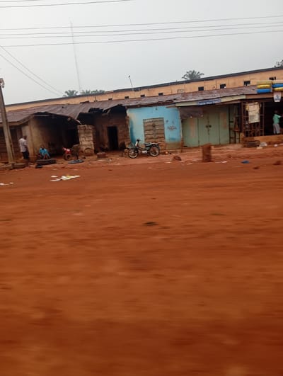 The image shows a street scene in Imilike, Nigeria. The foreground is dominated by a reddish-brown dirt road, with some scattered debris and a few rocks. In the mid-ground, a row of buildings with corrugated metal roofs lines the road. These buildings appear to be shops or small businesses, with some open doorways and shutters. One building is painted light blue, and another has green shutters. There are a few people visible in the scene. One person in a light-colored shirt and shorts stands near a doorway on the left. Another person in a blue shirt is sitting on a tire. Further to the right, a red and black motorcycle is parked, and another motorcycle with a white seat is visible near the blue building. In the background, a taller building with a light-colored facade and windows runs along the top of the scene. Overhead, power lines are visible against a hazy sky. A tall communication tower is also visible in the far distance, rising above the buildings and trees. The overall impression is one of a rural or semi-urban commercial area.