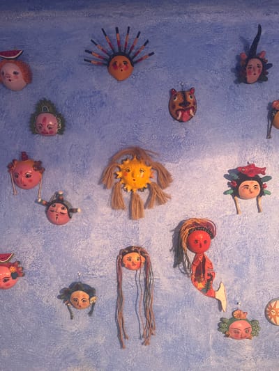 A collection of whimsical, handcrafted dolls or ornaments decorates a textured, light-blue wall. The objects include stylized faces with varying features and adornments like yarn hair, wooden spikes, and decorative embellishments. Some figures have distinct shapes, like a sun with yarn rays or a mermaid with a painted tail. The diverse range of designs creates an imaginative, folk-art aesthetic.