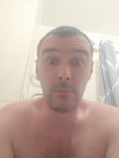 The image shows a close-up, upside-down selfie of a person in what appears to be a bathroom. The subject is a fair-skinned male with short, dark hair and a light beard. His eyes are visible, and he has a neutral expression. The background consists of white tiled walls and a shower curtain with a grey and white abstract pattern. The lighting is bright, suggesting daytime, and there are no obvious weather cues. The presence of tiles and a shower curtain strongly indicates the setting is a bathroom, possibly a shower, due to the slightly hazy appearance which might be steam. The image is a personal photograph, likely taken by the subject himself. No text is visible in the image.