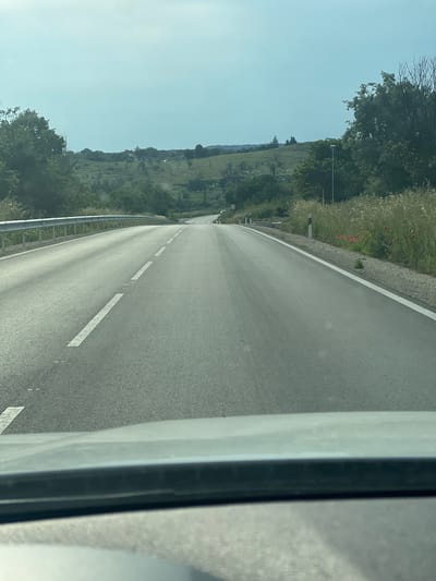 The image depicts a rural or semi-rural road scene in Tsarkva, Bulgaria, captured from the interior of a vehicle looking forward. The bottom portion of the image shows the top of a vehicle's dashboard or windshield frame.
The asphalt road, marked with a dashed white centerline and solid white shoulder lines, appears to ascend gently and curve slightly into the distance. The road surface is grey and shows minor imperfections.
To the left of the road, a silver metal guardrail runs parallel to the pavement. Beyond the guardrail, dense green foliage gives way to rolling hills covered in lush green grass and scattered trees, predominantly deciduous. Several small, light-colored buildings are visible in the mid-ground on these hills.
To the right of the road, a strip of tall, dry, light-brown grass and weeds lines the shoulder. Within this vegetation, several clusters of red poppies are visible. White and red striped marker posts are spaced along the roadside. Further back, green trees and shrubs are present, including a slender utility pole topped with a black fixture. Similar green hills with distant structures are visible beyond the immediate roadside vegetation.
The sky is a pale blue, appearing hazy and bright, indicating daytime conditions. No people or animals are visible in the scene.