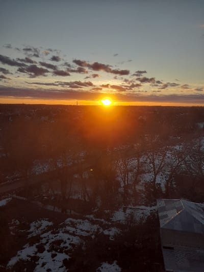 This image presents an elevated, panoramic view of a landscape identified as Borodianka, Ukraine, captured during either sunrise or sunset. The primary light source is a intensely bright, orange-yellow sun situated low on the horizon, slightly right of the frame's center, emitting significant glare. The sky transitions from a light blue overhead to warm orange and yellow tones near the horizon, populated by scattered dark-bottomed clouds illuminated from beneath. The distant horizon reveals a low-profile urban or town silhouette, with a slender, dark vertical structure visible to the left of the sun. The midground terrain is characterized by bare trees, indicative of winter or early spring conditions, interspersed with irregular patches of white snow covering the dark ground. Dark, unpaved-appearing paths are discernible winding through the foliage. In the bottom right foreground, a section of a multi-faceted roof, possibly constructed from corrugated metal or tiles, is partially visible. No individuals or specific actions are present in the scene.