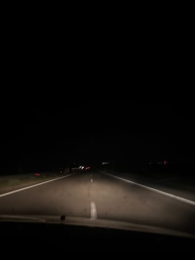The image depicts a nighttime view from the perspective of a vehicle traveling on a dark, asphalt road. The headlights of the vehicle illuminate the road ahead, revealing white lane markings. In the distance, faint streaks of light suggest other vehicles on the road. The sky is completely black, indicating it is nighttime. The immediate foreground shows a blurry portion of the vehicle's interior, likely the dashboard or windshield. The overall scene is dark and gives a sense of movement. There is no specific indication of the city of Vratitsa, Bulgaria, within the image itself.