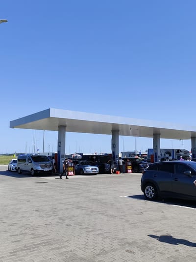 The image depicts a sunny day at a gas station in Chomentowo, Poland. The primary subject is a covered refueling area with multiple vehicles and people present. To the left, a silver van with license plate "JVV 543" is parked. Next to it, a white car is also parked. Under the canopy, several cars are being refueled or are waiting. A man in dark clothing and a face mask is walking away from a fuel pump. To the right of this man, an Audi sedan with license plate "HRC 60 333" is visible. Further to the right, a dark-colored SUV is parked. In the background, there are more fuel pumps, a large truck, and various structures associated with a gas station and its surroundings, including streetlights and a clear blue sky. The ground is paved with interlocking grey bricks.