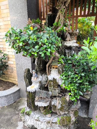 An outdoor ornamental rock garden and water feature is depicted in Thành phố Quảng Ngãi, Vietnam. The multi-tiered structure is constructed from grey, moss-covered rocks with prominent white, textured mineral-like deposits, suggestive of a dormant or artificial waterfall system. Integrated plant life includes a prominent bonsai-style specimen with dense, dark green foliage on the left, and various smaller vines and plants growing throughout the rock formations. Two figurines are positioned on the rockwork: an elderly man with a beard, dressed in robes, seated at a small table with cups on an upper tier, and a man wearing a conical hat, seated with a fishing rod on a lower tier to the left. A miniature wooden hut and a small pagoda are also nestled within the display. The immediate ground consists of grey concrete with a curved grey granite curb. Adjacent to the display on the left are a light-colored brick wall and a dark grey speckled pillar. In the background, a brown wooden lattice screen is visible, beyond which lush green foliage indicates an outdoor garden setting.