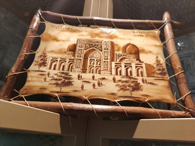 A close-up, angled shot shows a framed artwork featuring a depiction of the Tashkent, Uzbekistan skyline. The artwork is made of leather and stretched across a wooden frame constructed from rough, bark-covered branches secured with twine. The image on the leather displays architectural structures with domes and intricate geometric patterns, reminiscent of Central Asian mosques or madrasas. Small trees and figures of people are also visible in the scene, rendered in a brown, sepia-toned style. The artwork is suspended by twine from a horizontal wooden beam. The background is blurred, showing a dark, textured surface and hints of vertical elements. The foreground consists of a light brown, textured surface. The artwork appears to be a souvenir or decorative piece.