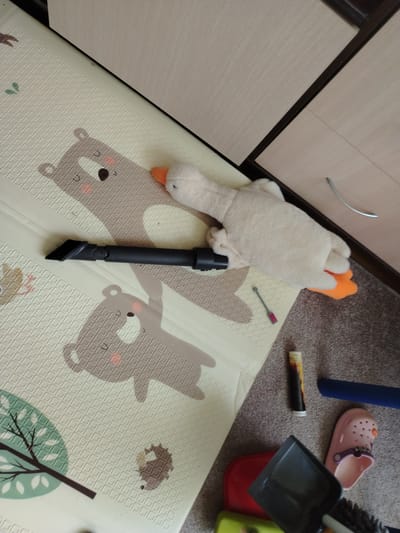 The image depicts an indoor scene, likely a room in a residential dwelling, located in the Тулаўскі сельскі Савет, Belarus. A light-colored play mat with printed animal designs, including bears and a hedgehog, covers a portion of the floor. A plush toy duck, predominantly white with an orange beak and feet, lies on the mat. A black vacuum cleaner attachment with a brush head is positioned on the mat, adjacent to the duck. In the background, a wooden cabinet with light-colored drawers is visible. On the carpeted floor to the right of the mat, several items are scattered: a pink clog-style sandal, a dustpan, a red object that appears to be a toy, a green object, a cylindrical tube with a dark patterned label, and a dark blue cylindrical object. The lighting suggests an indoor setting, possibly during daylight hours, with shadows cast by the objects.