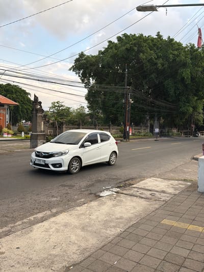 A white Honda Brio is driving on a street in Kubutambahan, Indonesia. The car is in the foreground, on the left side of the road, facing away from the camera. The street is paved with asphalt and has yellow lane markings. On the right side of the street, there is a sidewalk with paving stones and a yellow marking. A large, mature tree with dense green foliage dominates the background on the right side of the image. A street light hangs over the road on the left. In the far background, there are traditional Balinese stone gates and buildings with orange roofs. The sky is overcast with some blue patches.