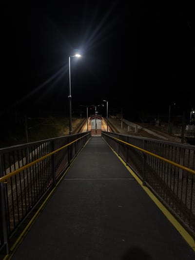 A ground-level, night-time, eye-level shot depicts a pedestrian overpass at a train station, identified as Bassendean, Australia, based on location metadata. The overpass spans a train track, visible between the overpass structure. The overpass has dark pavement and yellow railings on both sides. A lamppost on the left shines brightly. In the background, at the end of the overpass, are train station structures. The surrounding environment is dark, and details are difficult to distinguish due to the low light conditions. No people are clearly visible in the image.