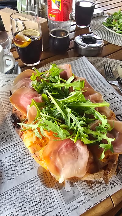 The image captures an outdoor dining scene at an establishment in Asparuhovo, Bulgaria. A table is set with a pizza topped with prosciutto and arugula, served on a newspaper. A glass of dark liquid with lemon slices and ice sits to the left, accompanied by a small glass and a white mug. To the right, a glass of dark beverage, a bottle of Coca-Cola, and an ashtray are visible. A plate of salad and cutlery complete the setting. The background suggests an urban outdoor environment.