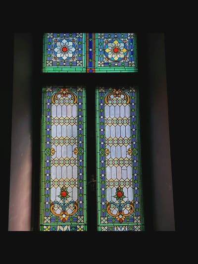 The image shows a close-up view of a stained-glass window, likely located in Saint-Août, France. The window is divided into three sections: a horizontal rectangular section at the top, and two tall, vertical rectangular sections below. The top section features intricate geometric patterns and floral motifs in shades of blue, green, yellow, and red, with a central circular design containing a red starburst. The two lower sections are identical, each depicting a tall, slender design with repeating geometric patterns of circles and diamond shapes in shades of white, yellow, pink, blue, and green. Each lower panel is framed by a wider border of green, blue, and purple glass. At the bottom of each lower panel, there is a detailed floral design with a prominent red rosebud atop a stem, flanked by stylized leaves and decorative scrollwork in orange and yellow. The overall impression is one of elaborate craftsmanship and vibrant color. The window appears to be set within a dark, possibly interior, space, with the light filtering through the stained glass casting soft patterns. The provided crops highlight specific sections of the stained glass, emphasizing the detailed patterns and motifs.