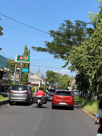 The image depicts a street scene in Pagerwojo, Indonesia, under a clear blue sky. A two-lane asphalt road is visible, with traffic moving in the same direction. In the foreground, the rear of a silver SUV is prominent, displaying a license plate ending in "N". Beside it, a motorcycle rider in a red jacket is moving forward, equipped with a large storage box attached to the back. Further ahead, an orange hatchback is visible, its license plate ending in "BBR".
On the left side of the road, a pole displays a prominent "K24" sign, indicating a pharmacy named "Apotek K24" with the slogan "Apoteknya Indonesia SEDIA SUSU 24 JAM". Below it, a smaller blue sign reads "WEDRINK". Adjacent to the pharmacy are various buildings and shops, some with awnings and parked motorcycles. Lush green trees line both sides of the street, with dense foliage overhead partially obscuring the sky. Telephone wires crisscross above the road. The overall impression is of a bustling urban environment on a sunny day.