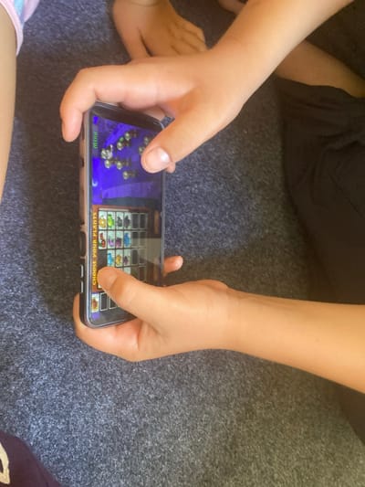 A young child's hands are holding a smartphone, displaying a game with a purple background and various plant icons. The game screen shows a grid with a vertical text overlay reading "CHOOSE YOUR PLANTS". The child's right index finger is pressing on one of the plant icons in the grid. The smartphone is a dark color with a silver frame. The child is sitting on a dark grey, textured surface, possibly a carpet or rug. Another child's leg, wearing pink and white patterned fabric, is visible on the left side of the frame. The lighting suggests an indoor environment, possibly during daylight hours. The scene does not contain any recognizable landmarks or features that would allow for localization to Arbagish, Uzbekistan.