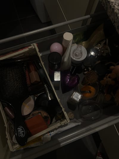 The image shows the inside of an open drawer in a bathroom, located at 56.049, 9.926, Skanderborg, Denmark. The drawer is filled with a variety of personal care items, including hairbrushes, makeup, toiletries, and a decorative object resembling a dried flower arrangement. Most of the items are concealed by darkness but some are visible, such as a hairbrush with the word 'BRON' printed on it, a makeup compact, and a tube of lotion labeled 'Wet Brush'. The drawer's contents are scattered and chaotic, indicating that it is used regularly and is not organized. The drawer is made of metal and appears to be part of a larger cabinet. The surrounding area is dim and the image appears to be taken from a slightly overhead angle.