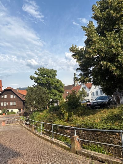 This is a medium shot, outdoor, vertical photograph taken in Gernsbach, Germany. The image captures a street scene with a focus on a cobbled pathway leading upwards alongside a stone wall with a metal railing. Dense green foliage and trees line the wall, partially obscuring buildings in the background.
In the background, several buildings are visible. One is a dark wooden house with multiple windows and a pitched, tiled roof. Adjacent to it, a lighter-colored building with white shutters and a tiled roof also stands. Further back, the steeple of a church or tower can be seen rising above the trees. A dark-colored car is parked on the street behind the railing, with its license plate partially visible, reading "RA-RR 813".
The sky above is a mix of blue and white clouds, suggesting it is daytime, likely late morning or afternoon, with fair weather. The sun is casting shadows, indicating a bright day. The overall atmosphere is calm and depicts a quaint European town. There are no people visible in the frame. The scene is tranquil and offers a glimpse into a residential or historic area of the town.