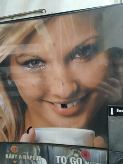 The media file is a photograph, likely taken at a vending machine in Choceň, Czechia. The foreground features a close-up of a poster, which is likely advertising coffee or beverages. The poster depicts a woman's face with blonde hair and striking eye makeup. Her mouth is slightly open, revealing a missing tooth. There are some smudges or dirt marks on her face, adding a grunge aesthetic. A finger is visible in the lower left corner, pointing towards the poster.

Below the poster, a section of the vending machine is visible, with text in Czech. This text includes "KÁVY A NÁPOJE" which translates to "COFFEES AND DRINKS", and "TO GO NA CESTU" which means "TO GO ON THE WAY". There is also a small circular icon with a coffee cup symbol. To the right of the poster, a black sign with a white arrow pointing down is visible, with the word "Škrá" partially visible, likely part of a word related to "scratch" or "scratch card" in Czech.

The overall impression is of an advertisement that has been potentially vandalized or aged, with the smudges and the missing tooth adding an unintentional, perhaps even unsettling, element to the image. The lighting suggests an indoor setting, typical of a vending machine area. No specific time of day or weather conditions are discernible. The emotion conveyed by the woman's expression is somewhat ambiguous due to the missing tooth and the smudges, but she appears to be looking directly at the viewer.