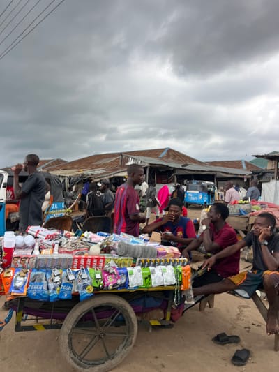 In Oko Amakom, Nigeria, under an overcast sky, a street market scene unfolds. The foreground features a cart laden with various goods, including bags of locks labeled "TIGGER" and "DORCO 4+1," as well as packaged accessories such as earphones and cables. Several young men are gathered around the cart and a nearby bench. One man in a black t-shirt and dark shorts, wearing a watch, is seated on the bench and talking on his phone. To his left, another young man in a maroon t-shirt is seated and looking down. In front of them, a young man in a red and blue striped t-shirt is seated and eating. Behind them, a man in a red and blue striped t-shirt is standing. Another man in a black t-shirt is standing to the left, holding a red object to his mouth. In the background, market stalls with corrugated metal roofs are visible, and a blue tricycle is parked. The ground is dirt.