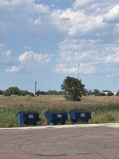 Three blue dumpsters, each with a black lid and bearing the "SCHABEN SANITATION" logo and phone number, are lined up in front of a tall green grassy area. The dumpsters are situated on a paved surface, possibly a parking lot. In the background, a field of dry, brown vegetation extends towards a line of trees and a distant tree with lush green foliage. A tall pole with a yellow and green sign is visible to the left of the tree, and a street light is positioned behind the tree on the right. The sky is a bright blue with scattered white clouds. The scene appears to be in an outdoor, semi-rural or commercial area, likely on the outskirts of North Platte, USA.