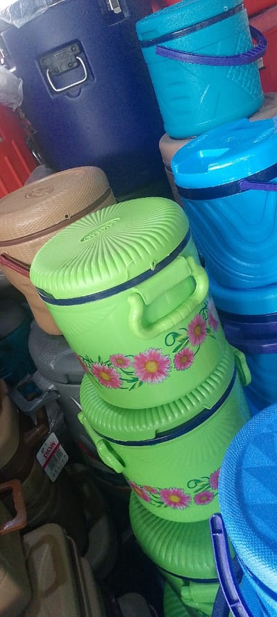 This image displays a crowded retail arrangement of various plastic household containers within a commercial establishment in Okere-Urhobo, Nigeria. The foreground and mid-ground are dominated by a stack of three lime-green plastic food warmers or coolers. The top two lime-green containers have side handles, dark blue bands below their ribbed lids, and feature a decal of pink daisies with green foliage. One lid bears the molded text "OMKAS." To the left, a beige plastic container with a woven texture and a red handle is visible. In the upper background, a large dark blue plastic cooler, partially covered by clear plastic, displays a silver-colored metal handle attached to a black rectangular base. Adjacent to it, a turquoise plastic bucket with a patterned surface and a purple handle is stacked. Additional brown and beige plastic items, including one with a "SALVIN" brand tag, are visible in the lower-left. No individuals are present, and no actions or interactions are depicted beyond the static arrangement of goods.