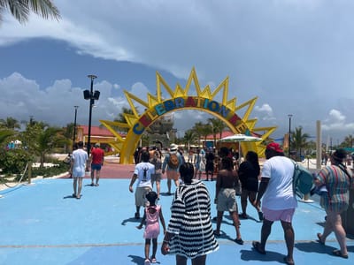 An overhead shot captures a group of people walking on a bright blue pavement towards a large, yellow sun-shaped archway with the word "CELEBRATION" in colorful block letters. The archway is adorned with lights and leads to a castle-like structure. Palm trees and a few buildings with red roofs are visible in the background, under a partly cloudy sky. The scene appears to be a public space, possibly a theme park or a cruise port, given the festive atmosphere and attire of the people. This location is consistent with a part of City of Freeport, Bahamas, known for its tourist attractions and cruise terminals.