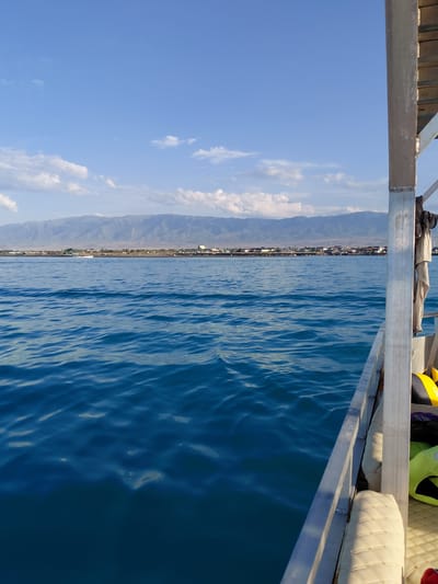 The image captures a view from a boat on the water, looking towards a city nestled at the base of a mountain range. The water is a deep blue with gentle ripples, reflecting the clear sky above. In the distance, Akshi, Kazakhstan, is visible as a line of buildings and structures along the shoreline, with a prominent mountain range forming a dramatic backdrop. The mountains appear arid and rugged, with a lighter tone than the water. The sky is a brilliant blue, dotted with scattered white clouds. The right side of the image shows a portion of the boat's railing and some seating, along with a yellow life preserver, suggesting recreational activity. A small white boat is visible in the distance on the water, near the shore. The overall scene is one of a serene coastal landscape with a distant urban presence.