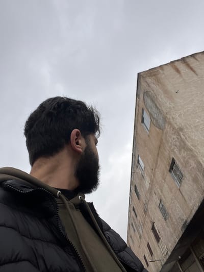 The image depicts a scene in Dahiyat Harasta, Syria, under an overcast sky. A male individual, visible from the chest up and positioned on the left side of the frame, is observed from a slightly rear-left perspective. He has dark, wavy hair and a full beard. The individual is wearing a black puffer jacket over an olive-green hooded sweatshirt, with the hood's drawstrings visible. His head is turned to the right, with his gaze directed upwards towards a multi-story building.
The building occupies the right side of the frame, angled upwards and right, and appears to be constructed of faded, light beige or dirty cream-colored concrete or stucco, showing signs of significant weathering and discoloration. Multiple rectangular windows are visible on its facade, some appearing broken or in disrepair, others with visible frames. A prominent, large, oval-shaped discolored patch is present on the upper portion of the building's facade. A small, dark, bird-like object is discernable in the sky near the upper right corner of the building. The sky is a uniform light grey, indicating an overcast condition. The overall environment presents as an urban setting with aging infrastructure.