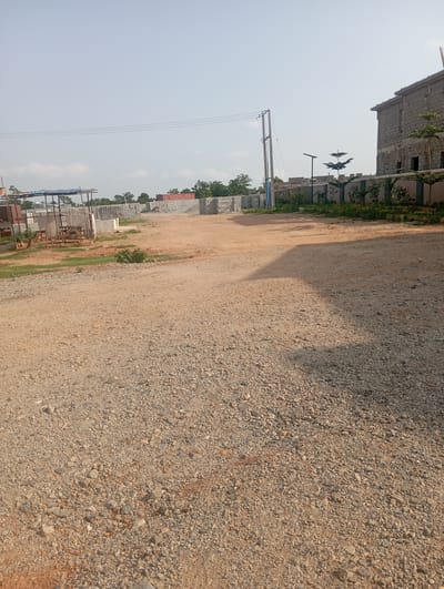 The image depicts an outdoor scene in Tasha, Nigeria. The foreground is a gravel road. In the middle ground, there is a construction site with stacks of concrete blocks and what appears to be a partially constructed building made of concrete blocks. A power pole with electrical wires is visible on the left. In the background, more construction materials are present, including what looks like shipping containers. A wall with decorative fencing and small trees is also visible. The sky is clear and blue. There are no people visible in the image.