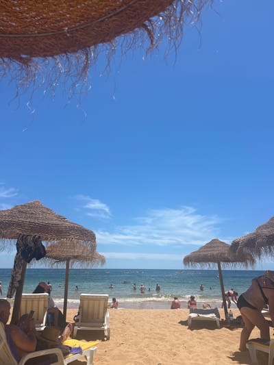 The image captures a sun-drenched beach scene at Praia Dona Ana in Lagos, Portugal. The foreground is dominated by sandy beach with several empty and occupied lounge chairs, some shaded by thatched umbrellas. In the midground, people are scattered across the beach and wading in the clear, blue ocean. The water is calm with gentle waves, and several individuals are visible swimming or standing in the sea. In the distance, more people can be seen enjoying the water, with a few small boats or paddleboards present. The sky is a vibrant blue with scattered wispy white clouds, indicating a bright, sunny day. The overall atmosphere is one of leisure and relaxation, typical of a popular beach destination.